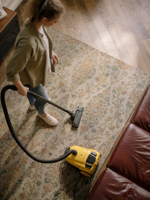 A professional cleaning technician dressed in a full white protective suit, gloves, and face mask is operating a steam cleaner on a beige carpet in a bright, modern living room. The room features a la