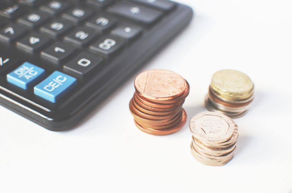 Close-up image showing a black calculator with blue and white buttons in the top left corner, next to three neatly stacked piles of coins of various denominations, including pennies and larger coins, 