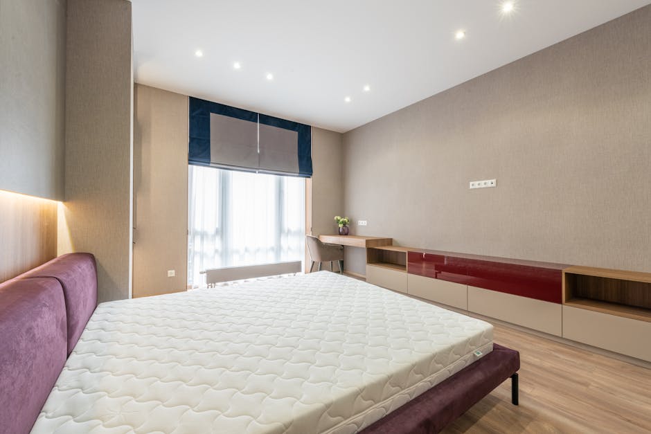 A modern bedroom showcasing a large, unmade bed with a white mattress and a purple upholstered headboard, positioned against a wall with beige textured wallpaper. The room features a wooden floor and a built-in low cabinet with a mix of open shelves and closed drawers in a beige and dark red finish, running along the right side wall. To the left, a window with sheer white curtains and a blue roman blind allows natural light into the room, illuminating the simple decor. A small desk with a chair and a potted plant is situated near the window, and the ceiling is fitted with multiple recessed lights, creating bright, even lighting. The overall appearance is clean and tidy, reflecting professional domestic cleaning standards in line with surface cleaning and hygiene practices by Merton Carpet Cleaners.