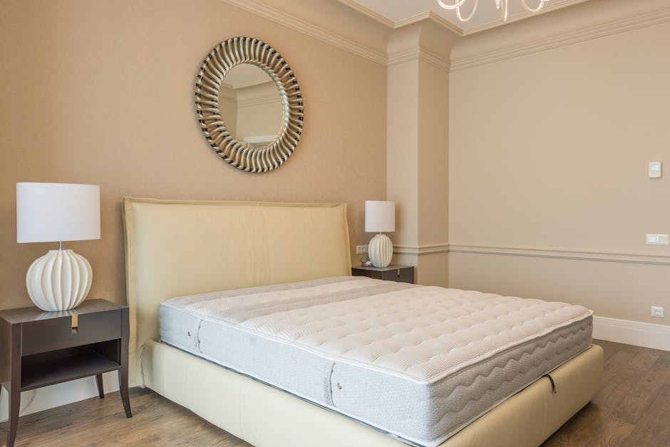 A modern bedroom featuring a white quilted mattress on a bed with a light beige upholstered headboard, situated against a dark wood paneled accent wall. On either side of the bed, there are black wall-mounted reading lamps and matching round black bedside tables. To the left, a black metal shelving unit with multiple shelves is visible, and to the right, a white door is partially seen. The room is well-lit with overhead lighting, and the surface cleaning and sanitisation of the room suggest a professional domestic cleaning service, as promoted by Merton Carpet Cleaners on their website for surface cleaning and deep cleaning purposes.